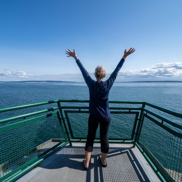 Woman passenger on the Port Townsend Ferry in Washington State stands with her arms up and back to the camera as the boat sails across Puget Sound